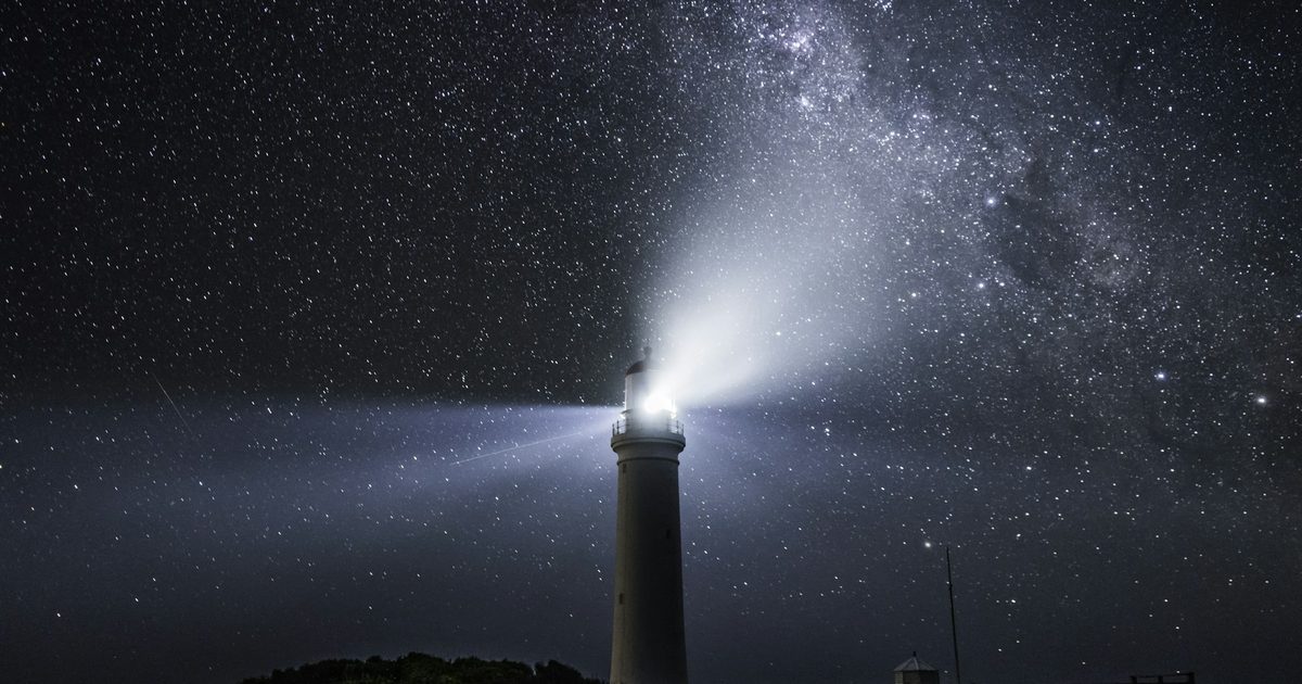 A lighthouse casting its beam across the sky beneath the Milky Way